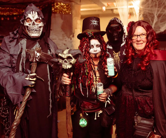 Group of four in Halloween costumes at an indoor party: skeletal grim reaper with scythe, voodoo-style witch with skull-topped staff and top hat, masked hooded figure, and a woman in a crimson wig and gothic cloak holding drinks.