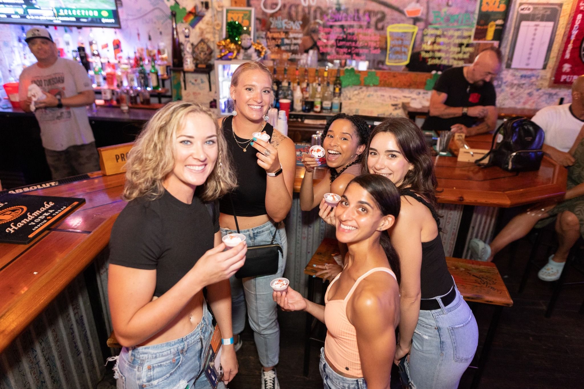 Five friends smiling and posing at a lively indoor bar counter, each holding a small shot cup, with bartenders, liquor bottles and a colorful chalkboard menu in the background — fun nightlife scene.