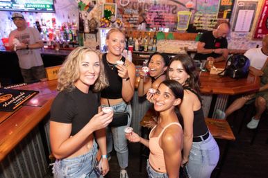 Five friends smiling and posing at a lively indoor bar counter, each holding a small shot cup, with bartenders, liquor bottles and a colorful chalkboard menu in the background — fun nightlife scene.
