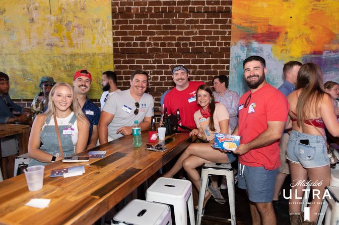 Smiling group of young adults at a lively urban bar with exposed brick and colorful murals, gathered around a wooden high-top table with beer bottles and a box of Twinkies.