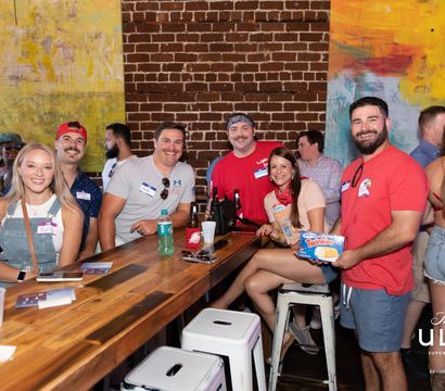 Smiling group of young adults at a lively urban bar with exposed brick and colorful murals, gathered around a wooden high-top table with beer bottles and a box of Twinkies.