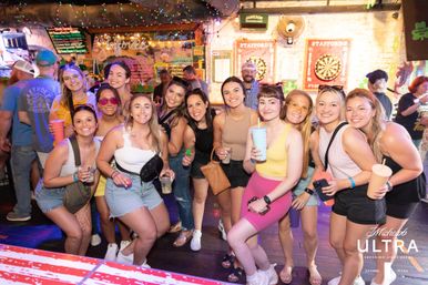Smiling group of young women posing with drinks at a lively neon-lit indoor bar and pub, dartboards on the wall and a crowded nightlife scene behind them.