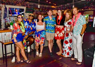 Seven friends in colorful Hawaiian shirts, floral dresses and leis smiling and posing inside a tropical-themed bar with string lights, a stocked counter and lively nightlife atmosphere