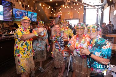 Group of adults in colorful Hawaiian shirts, leis, grass skirts and novelty tropical sunglasses raising drinks and cheering inside a lively indoor bar with hanging Edison bulbs and TV screens.