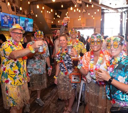 Group of adults in colorful Hawaiian shirts, leis, grass skirts and novelty tropical sunglasses raising drinks and cheering inside a lively indoor bar with hanging Edison bulbs and TV screens.