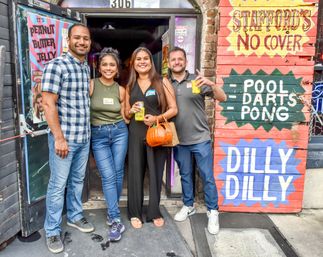 Four smiling friends posing at a colorful bar entrance with hand-painted signs for pool, darts and pong; two hold canned drinks and one carries a pumpkin-shaped purse, nightlife scene.