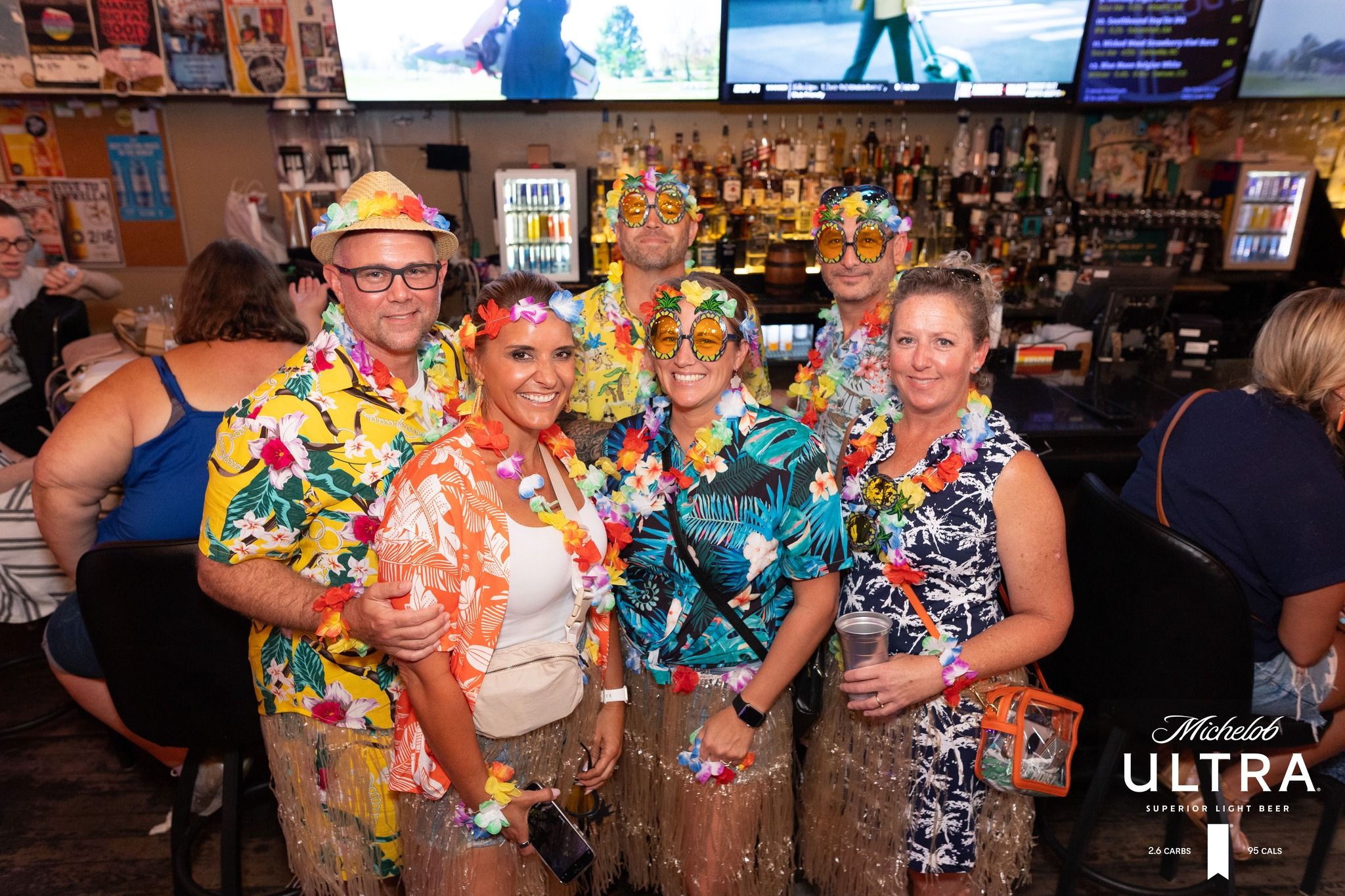 Six adults in colorful Hawaiian shirts, leis, grass skirts and novelty sunglasses smiling and posing at a lively indoor bar with TV screens and bottles behind them.