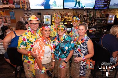 Six adults in colorful Hawaiian shirts, leis, grass skirts and novelty sunglasses smiling and posing at a lively indoor bar with TV screens and bottles behind them.