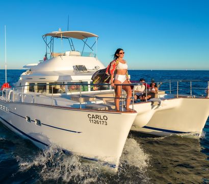 Sunlit white twin-hull motor yacht cruising on deep blue ocean at golden hour, people relaxing on the bow — woman in a white swimsuit stands at the rail and another in a white sundress poses at the front, with a rocky coastline and clear sky in the distance.