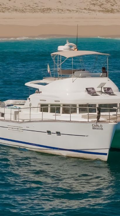 White catamaran anchored in turquoise coastal waters off a sandy beach, crew on deck and a small blue tender with passengers wearing orange life jackets.