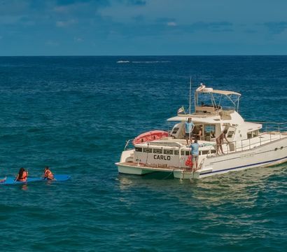 White motor yacht floating on deep-blue ocean under a sunny sky, passengers on deck and three people in orange life jackets paddling on a long blue board nearby.