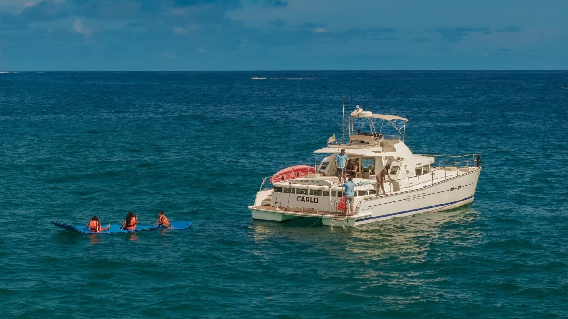 White motor yacht floating on deep-blue ocean under a sunny sky, passengers on deck and three people in orange life jackets paddling on a long blue board nearby.