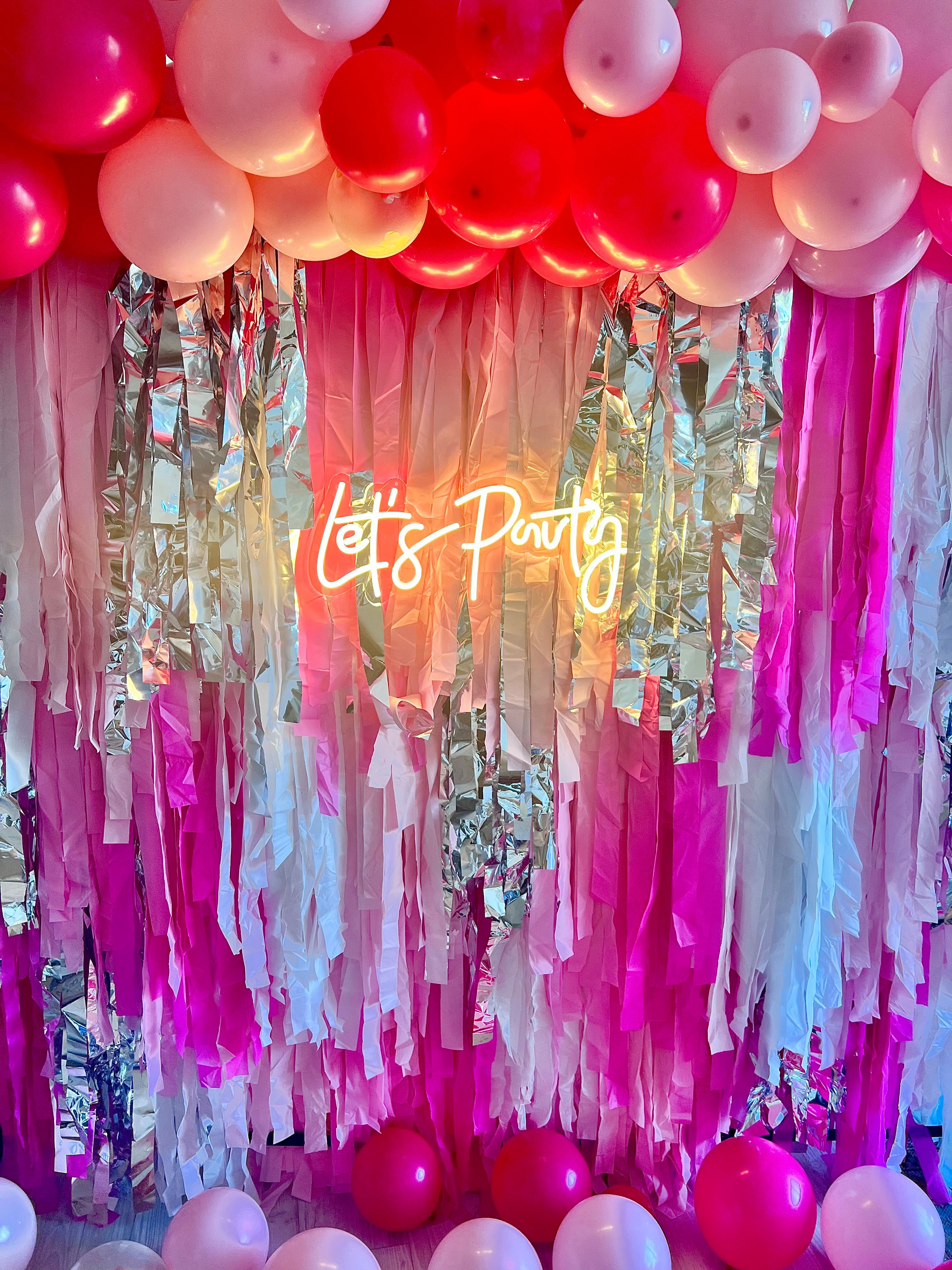 Festive indoor party backdrop with pink and red balloon garland, silver and ombré pink fringe streamers, glowing neon sign reading 'Let's Party', and scattered pink balloons on the floor.