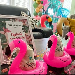 Tropical-themed indoor bachelorette party setup in a living room with a framed welcome sign, pink inflatable flamingo cup holders holding small favor bags, pastel balloon garland and palm-print backdrop on a table.