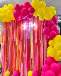 Vibrant indoor party backdrop with hot pink and sunny yellow balloon arch, long pink-orange streamers and glowing neon “Let's Party” sign for photo booth or event decor