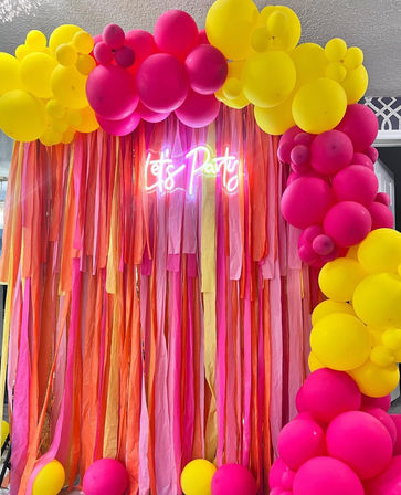 Vibrant indoor party backdrop with hot pink and sunny yellow balloon arch, long pink-orange streamers and glowing neon “Let's Party” sign for photo booth or event decor