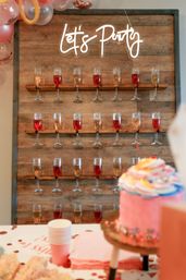 Wooden champagne wall displaying rows of wine flutes with red and sparkling drinks under a neon “Let’s Party” sign, pastel balloon garland at the top and a pink frosted cake with party cups in the foreground — festive party setup.