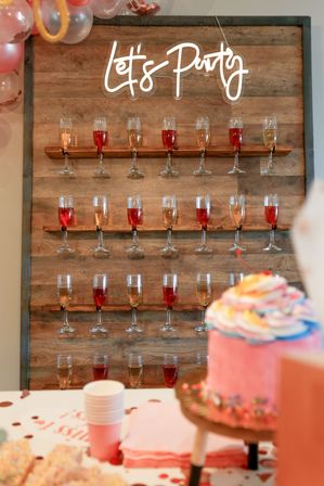 Indoor party decor: wooden champagne-glass wall with alternating pink and rosé drinks under a glowing “Let’s Party” sign, balloons and a pink-frosted cake with cups and napkins on the dessert table in front