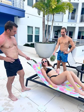 Poolside scene at a modern apartment courtyard — woman in a black bikini and heart-shaped sunglasses lounging on a colorful towel while two shirtless men in swim trunks playfully fan her with palm leaves near large planters and balcony apartments.