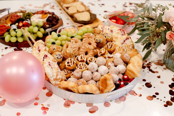 Festive brunch spread on a white table: assorted pastries (mini donuts, powdered donut holes, croissants, danishes), grapes and berries on a platter, pink balloon, rose bouquet and confetti