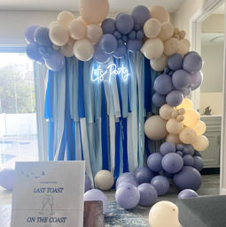 Coastal-inspired indoor party backdrop with blue ribbon curtain, lavender and cream balloon arch, scattered balloons on the floor and a glowing "Let's Party" neon sign in a home living room.