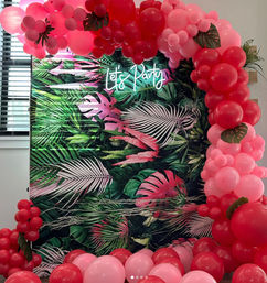 Indoor tropical party backdrop with green palm-leaf print, neon "Let’s Party" sign, and cascading pink and red balloon arch accented with faux monstera leaves