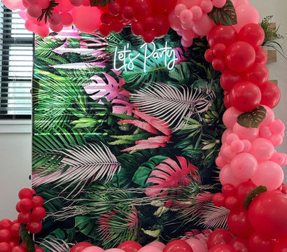 Indoor tropical party backdrop with green palm-leaf print, neon "Let’s Party" sign, and cascading pink and red balloon arch accented with faux monstera leaves