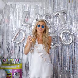 Smiling bride-to-be in a white dress and sash raising a champagne glass at an indoor bachelorette party with silver fringe backdrop, round sunglasses, and silver foil 'Last Fling' balloons