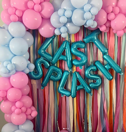 Playful pastel party backdrop with pink and baby-blue balloon garlands, teal foil letter balloons forming a celebration message, and multicolored pastel streamers for indoor party decor.