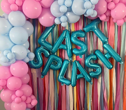 Playful pastel party backdrop with pink and baby-blue balloon garlands, teal foil letter balloons forming a celebration message, and multicolored pastel streamers for indoor party decor.