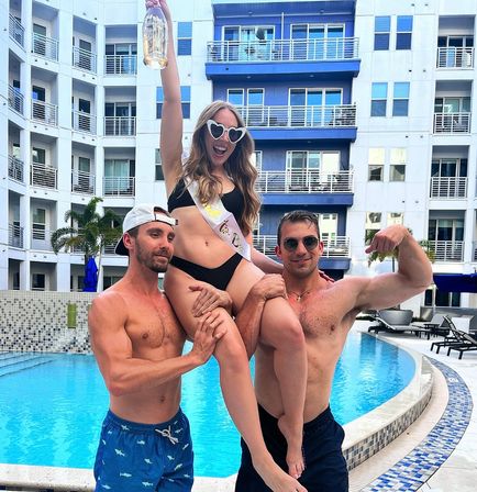 Bachelorette pool party: woman in black bikini and heart sunglasses wearing a sash and hoisting a champagne bottle while two shirtless men lift her at a city condo pool
