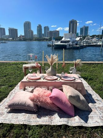 Cozy pink waterfront picnic tablescape with low table, plates, wine glasses, candles and pampas grass on rugs and pillows, overlooking a marina with yachts and a high-rise city skyline under a bright blue sky
