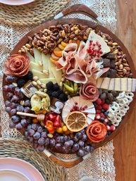 Rustic round charcuterie grazing board on lace placemat with grapes, assorted cheeses, prosciutto and salami roses, nuts, dried apricots, cornichons, honey and mixed berries.