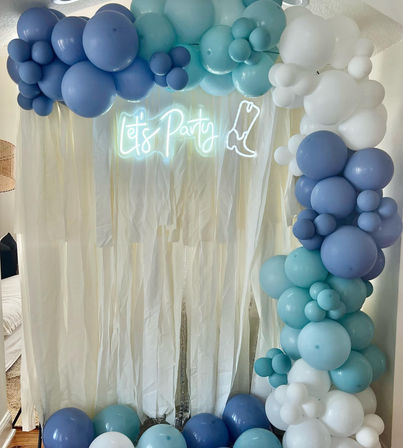Indoor photo booth backdrop featuring a blue, teal and white balloon arch, sheer white drapes, and a glowing neon “Let’s Party” sign with a boot outline.