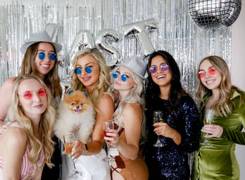 Party photo booth with six women in colorful round sunglasses and sparkly hats toasting with champagne in front of a silver tinsel backdrop and disco ball, one holding a fluffy Pomeranian