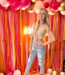 Smiling woman posing at an indoor party with pink, orange and gold balloons and streamers backdrop, wearing a gold crop top, ripped jeans and a celebratory sash.