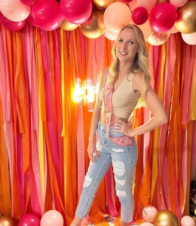 Smiling woman posing at an indoor party with pink, orange and gold balloons and streamers backdrop, wearing a gold crop top, ripped jeans and a celebratory sash.