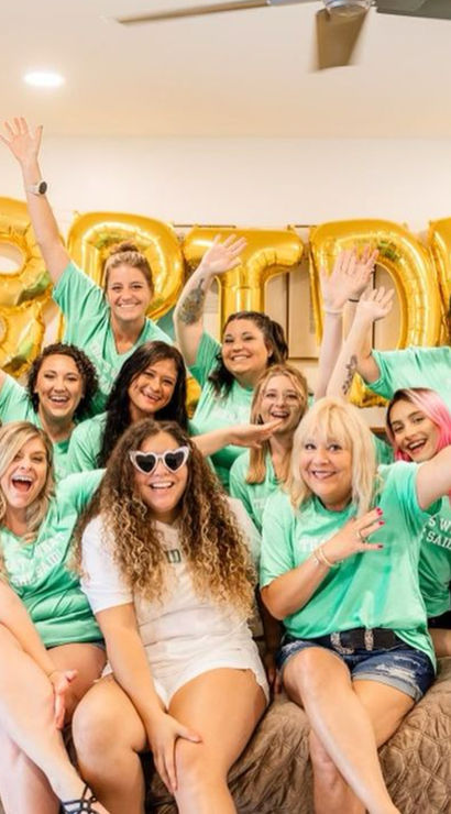 Cheerful bachelorette party group: women in matching teal shirts and denim shorts surround a bride in white with heart-shaped sunglasses, gold 'BRIDE' balloons on the wall in a bright indoor celebration.