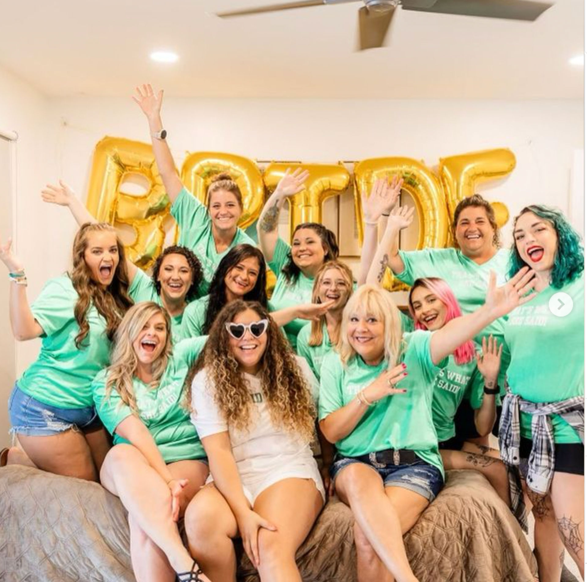Cheerful bachelorette party group: women in matching teal shirts and denim shorts surround a bride in white with heart-shaped sunglasses, gold 'BRIDE' balloons on the wall in a bright indoor celebration.