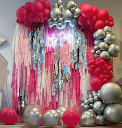 Pink and silver balloon arch framing metallic fringe curtain with neon 'Let's Party' sign — indoor photo-booth backdrop for birthdays and events.