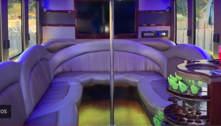 Party bus interior with purple LED lighting, wraparound gray leather seating, center dance pole, wall-mounted flat-screen TV and tiered cup holder station