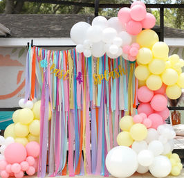 Outdoor pastel party photo booth with multicolored ribbon curtain, gold "splash" banner, and pastel pink, yellow and white balloon clusters framing the backdrop
