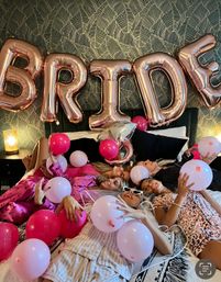 A group of friends in pajamas lounging on a bed for a bachelorette sleepover, surrounded by pink balloons and a rose-gold foil “BRIDE” banner against patterned wallpaper.