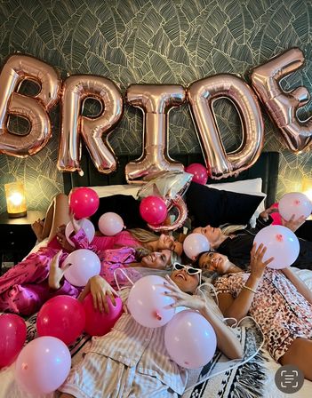 A group of friends in pajamas lounging on a bed for a bachelorette sleepover, surrounded by pink balloons and a rose-gold foil “BRIDE” banner against patterned wallpaper.