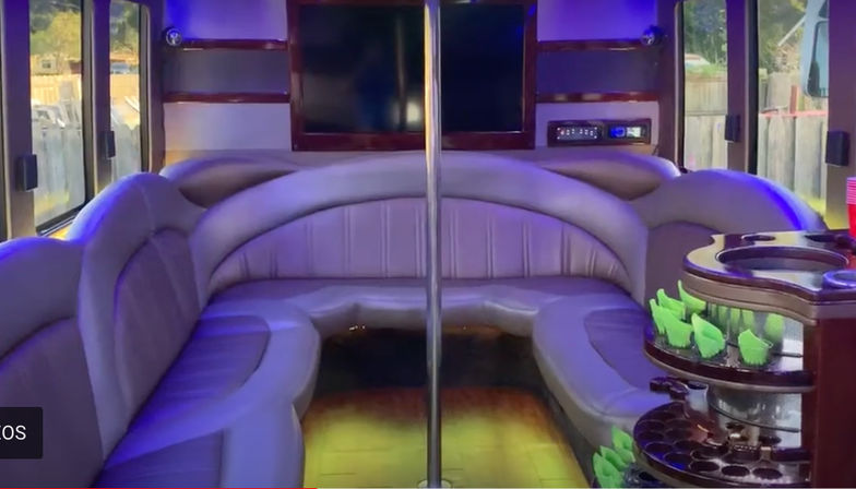 Party bus interior with curved gray leather lounge seating surrounding a central dance pole, purple LED lighting, wall-mounted TV, windows, and a rotating cup holder station with neon green cups.
