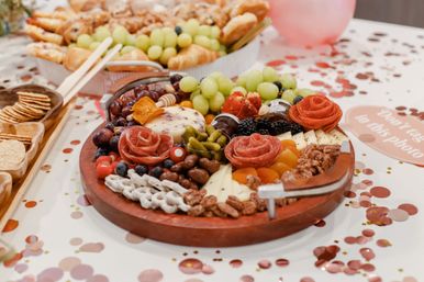 Colorful round charcuterie board on a confetti-covered party table with salami roses, assorted cheeses, grapes, blackberries, crackers and candied nuts.