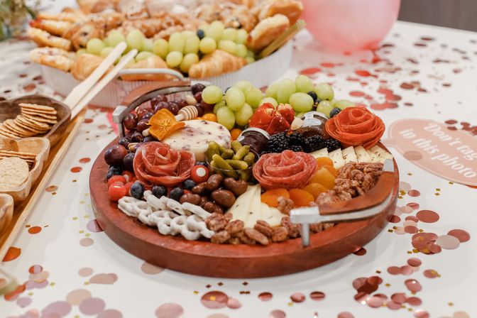 Colorful round charcuterie board on a confetti-covered party table with salami roses, assorted cheeses, grapes, blackberries, crackers and candied nuts.