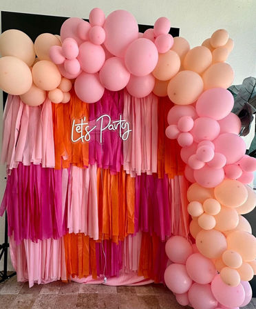 Vibrant pink and orange party photo backdrop with layered tassel fringe and pastel balloon garland arch, neon 'Let's Party' sign