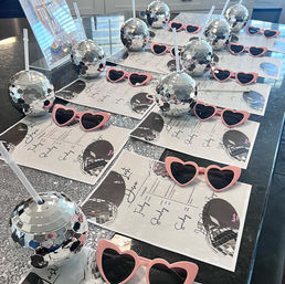 Disco-themed party favors: mirrored disco-ball tumblers with straws and pink heart-shaped sunglasses arranged on printed itineraries across a sparkly table or kitchen island.