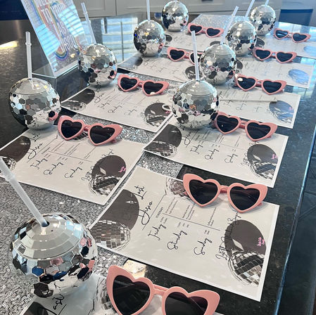 Disco-themed party favors: mirrored disco-ball tumblers with straws and pink heart-shaped sunglasses arranged on printed itineraries across a sparkly table or kitchen island.